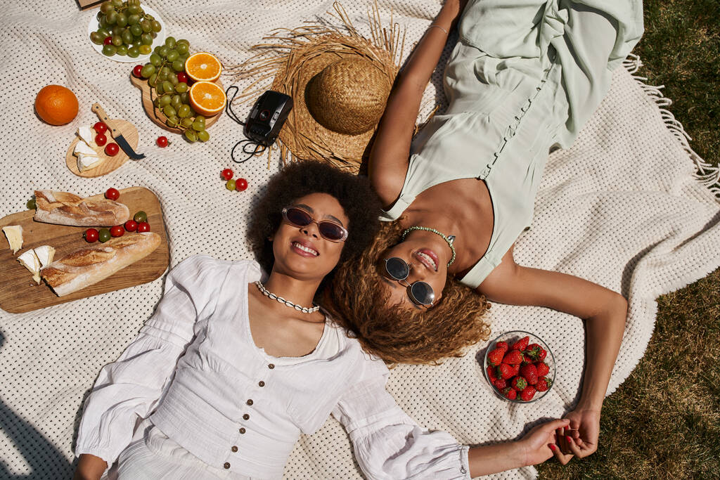 ξέγνοιαστες αφροαμερικανικές φίλες ξαπλωμένες σε κουβέρτα, φρούτα, λαχανικά, κορυφαία θέα, καλοκαιρινό πικ-νικ - Φωτογραφία, εικόνα