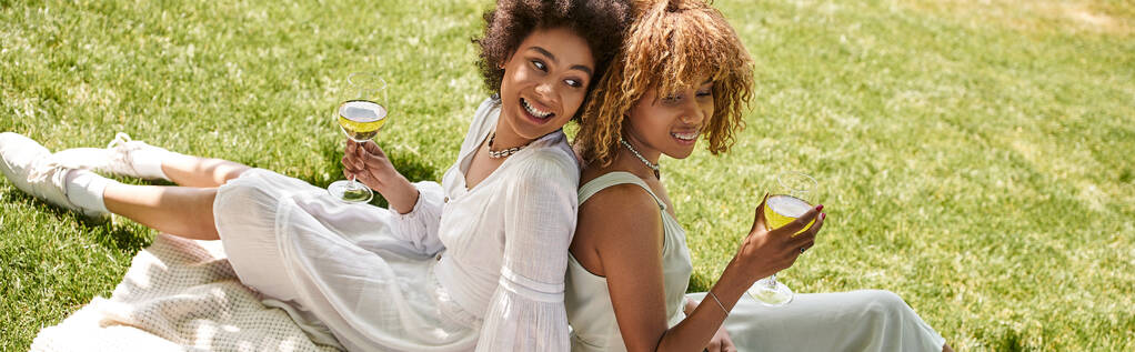 novias afroamericanas con copas de vino sentado espalda con espalda en el césped verde, pancarta - Foto, Imagen