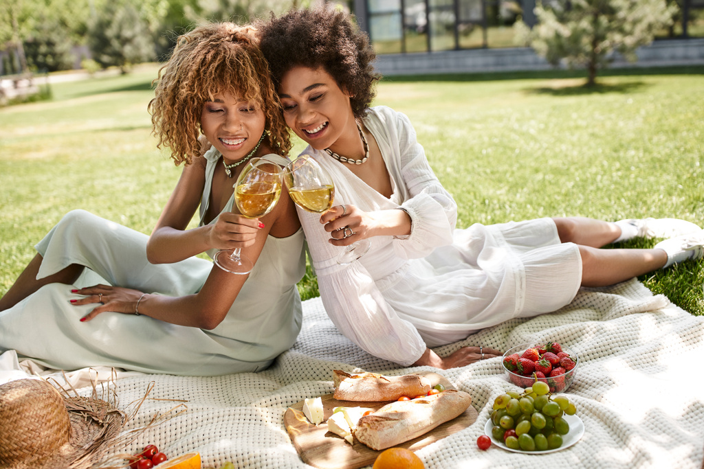 Afrykańskie amerykańskie dziewczyny siedzące z tyłu na plecach, brzęczące kieliszki do wina przy przekąskach, letni piknik - Zdjęcie, obraz