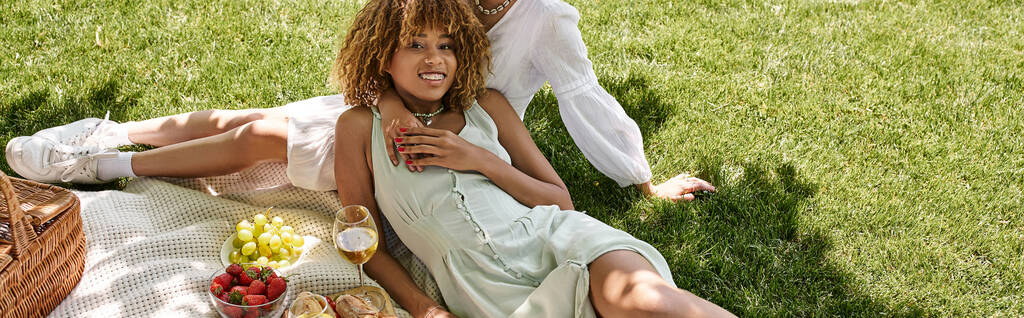 Kaygısız Afrikalı Amerikalı kadın kameraya bakıyor, kız arkadaşına yakın, parkta piknik, afiş - Fotoğraf, Görsel