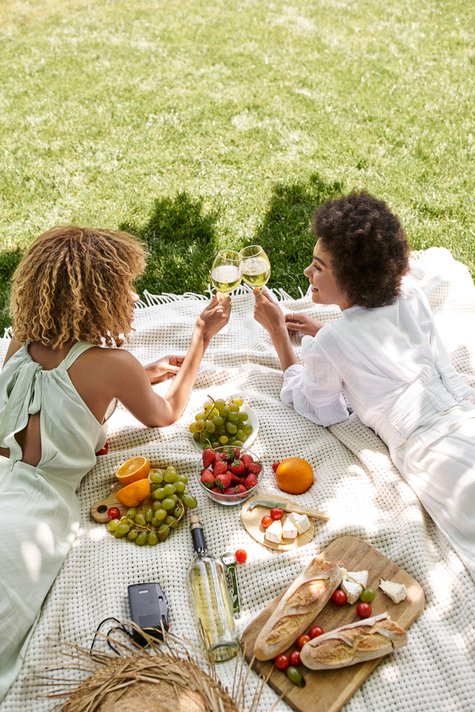 Afrikaans amerikaanse vriendinnen klinkende wijnglazen in de buurt van snacks op deken, zomer picknick - Foto, afbeelding