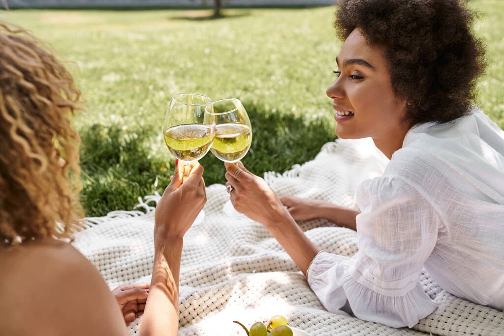 веселая африканская американка, звенящая бокалами вина с подружкой на одеяле во время пикника - Фото, изображение