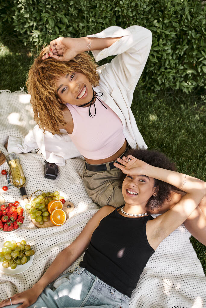 Kaygısız Afrikalı Amerikalı kız arkadaşların yaz pikniğinde şarap ve taze meyve yemesi. - Fotoğraf, Görsel