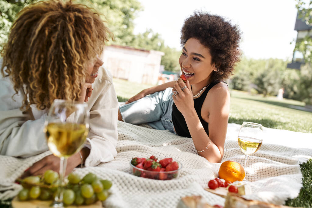 beztroska Afroamerykanka trzyma truskawkę i rozmawia z dziewczyną na letnim pikniku - Zdjęcie, obraz
