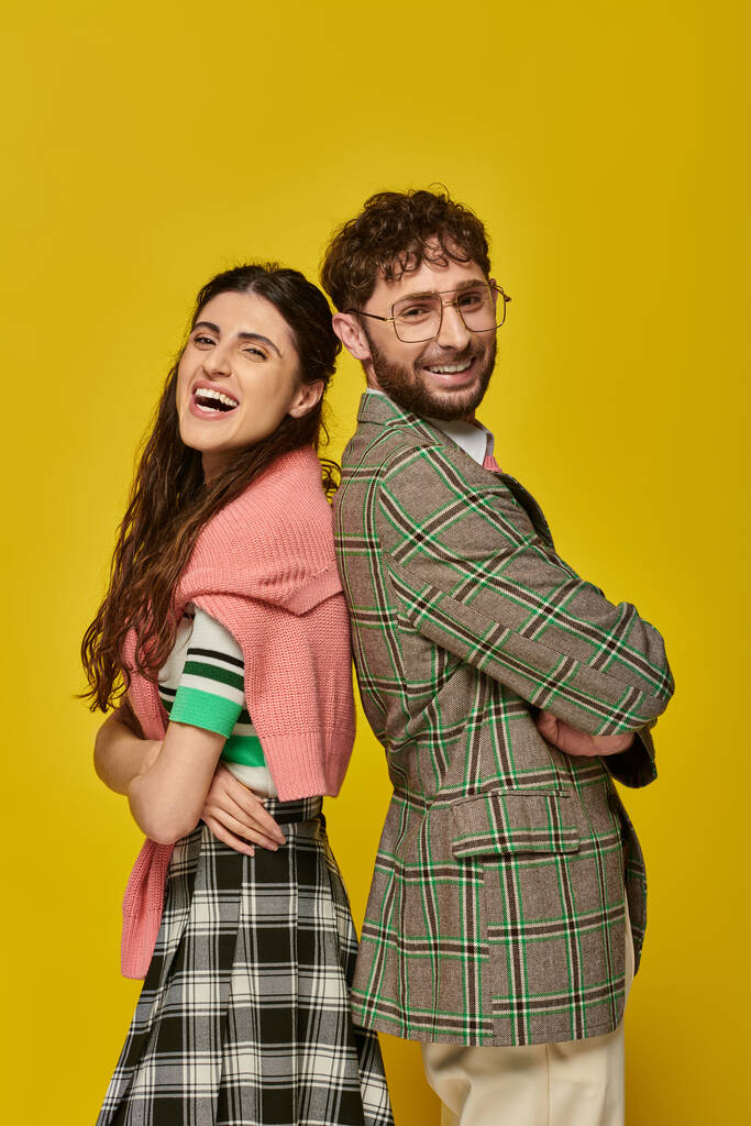 szczęśliwy stylowy mężczyzna i kobieta stojący plecami do siebie, pozowanie, żółte tło, strój studenta, śmiech - Zdjęcie, obraz