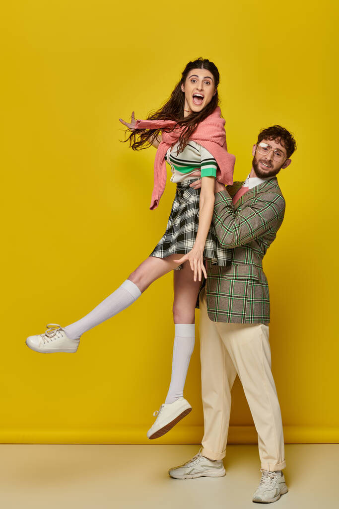 alegre casal se divertindo, homem de óculos levantando animado jovem mulher no fundo amarelo, estudantes - Foto, Imagem