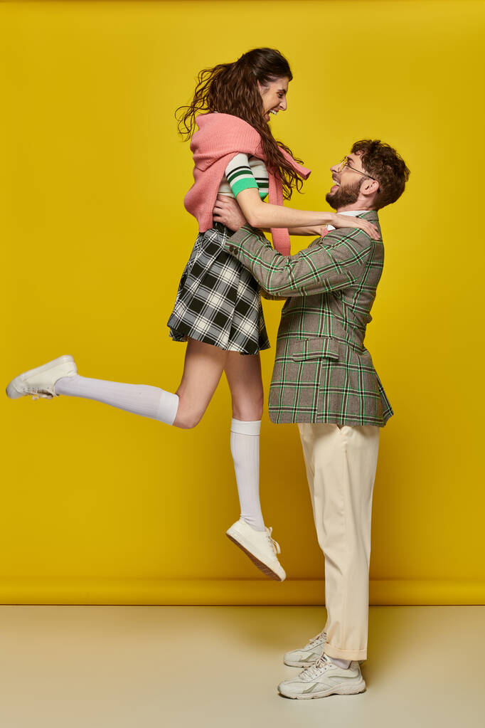 coppia gioiosa divertirsi, uomo in occhiali sollevamento eccitato giovane donna su sfondo giallo, studenti - Foto, immagini
