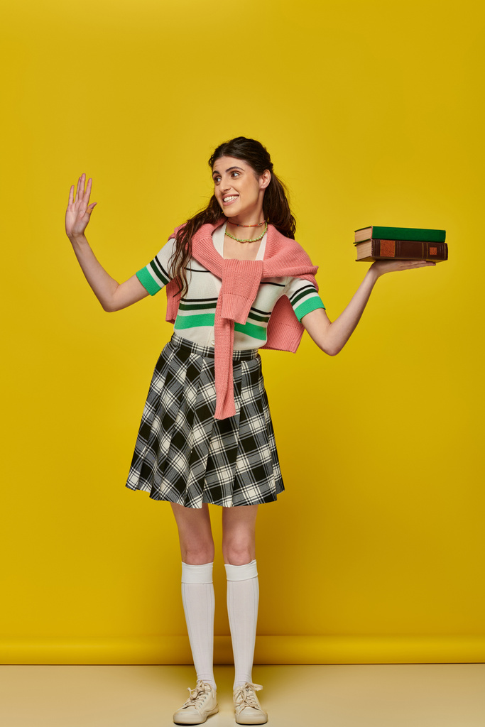 happy student with books, brunette woman in checkered skirt waving hand on yellow backdrop, brunette - Photo, Image