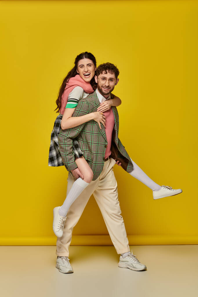 ενθουσιασμένοι ζευγάρι, γενειοφόρος νεαρός άνδρας piggybacking μελαχρινή γυναίκα σε κίτρινο φόντο, αστείοι μαθητές - Φωτογραφία, εικόνα