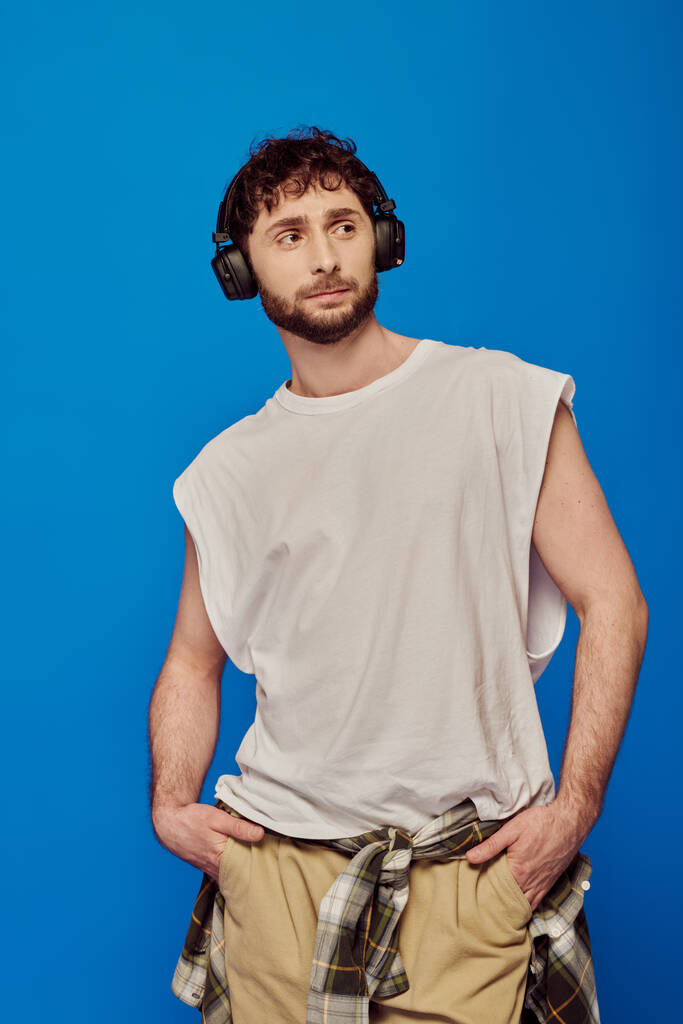 bearded man in wireless headphones listening music on blue background, white tank top, male fashion - Photo, Image