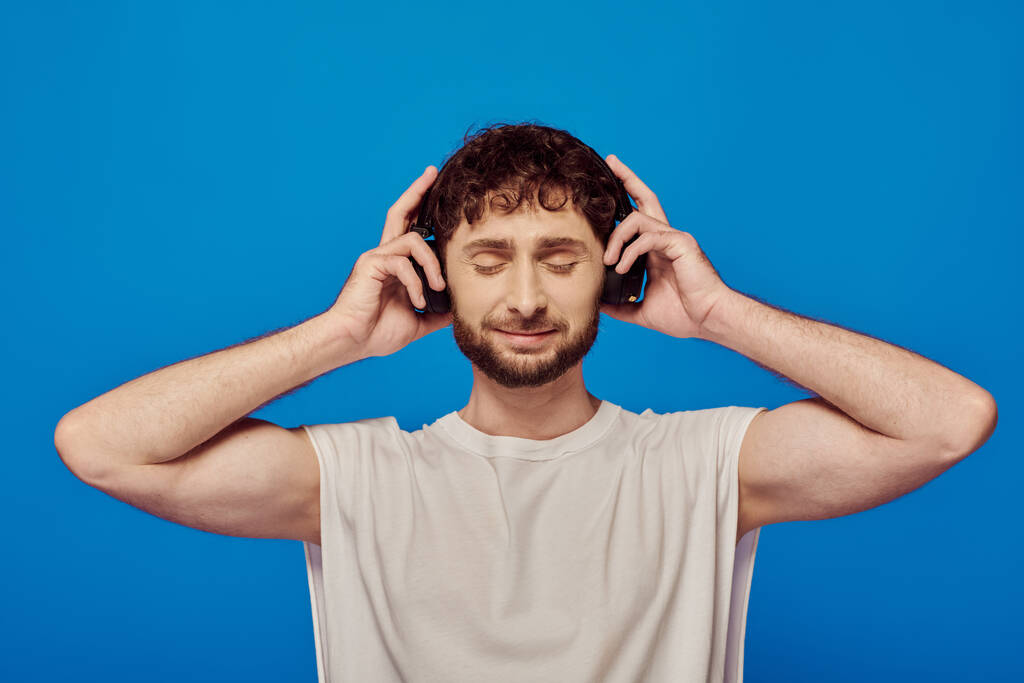 hombre complacido en auriculares inalámbricos escuchando música sobre fondo azul, camiseta blanca, ojos cerrados - Foto, Imagen