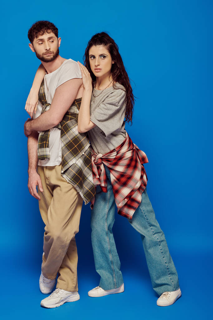 couple posing in street wear, blue backdrop, woman with bold makeup leaning on bearded man, trend - Photo, Image