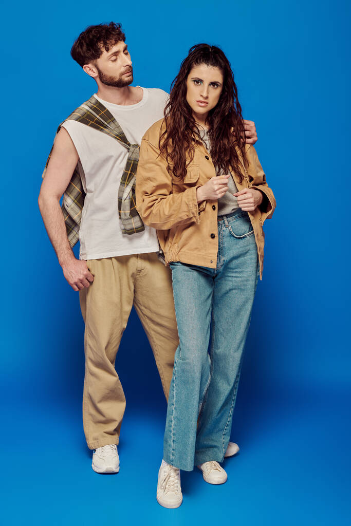 стильная пара позирует в повседневной одежде, синий фон, женщина стоит с бородатым мужчиной, смелый макияж - Фото, изображение
