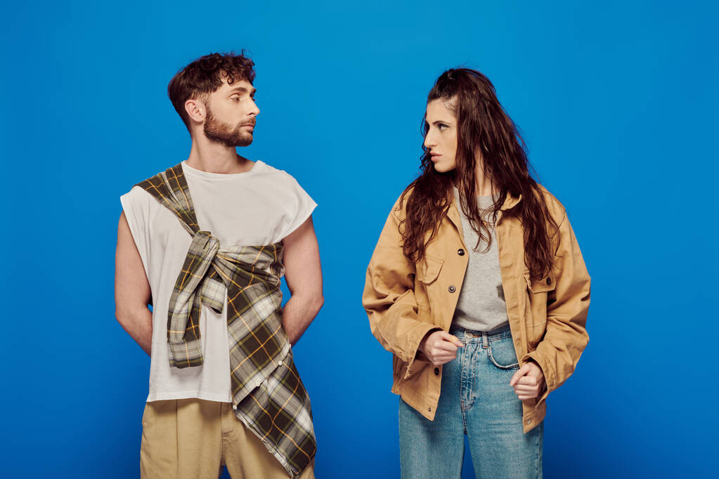 stylish couple in casual wear looking at each other, blue backdrop, woman standing with bearded man - Photo, Image