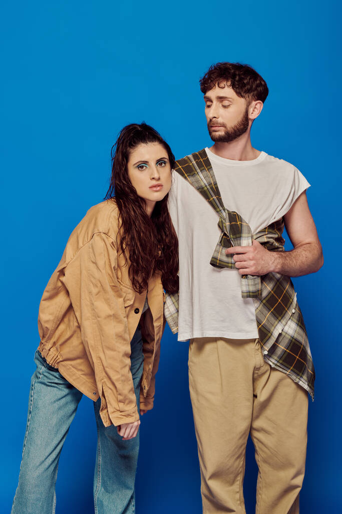 fashionable couple in casual wear posing on blue backdrop, bearded man hugging woman, bold makeup - Photo, Image