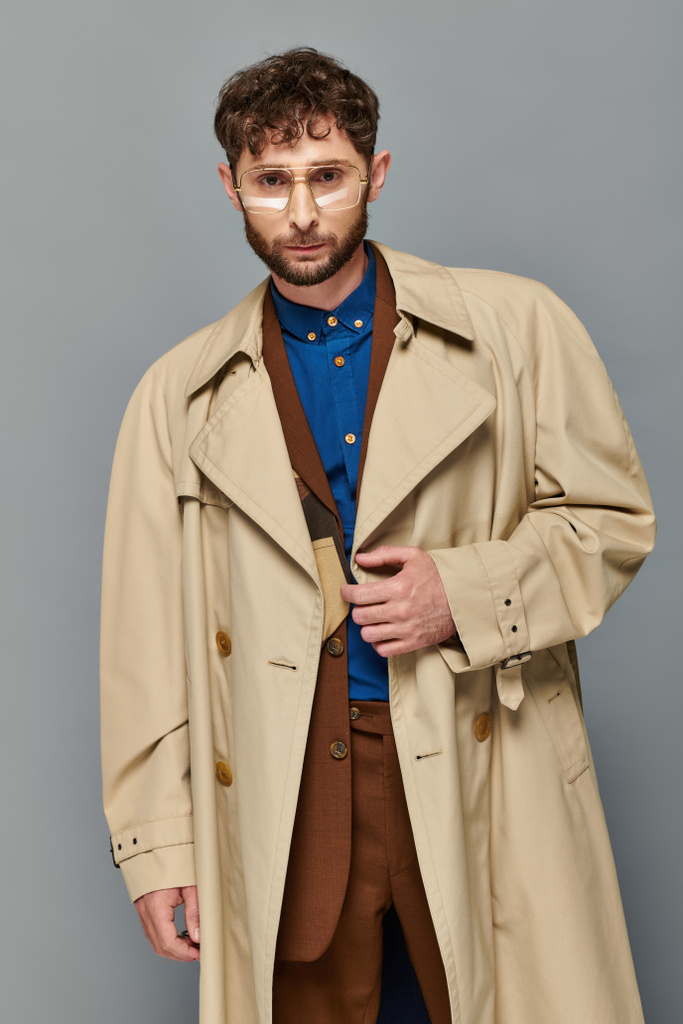 outerwear, trendy man in glasses and trench coat posing on grey backdrop, cozy layers, fall fashion - Photo, Image