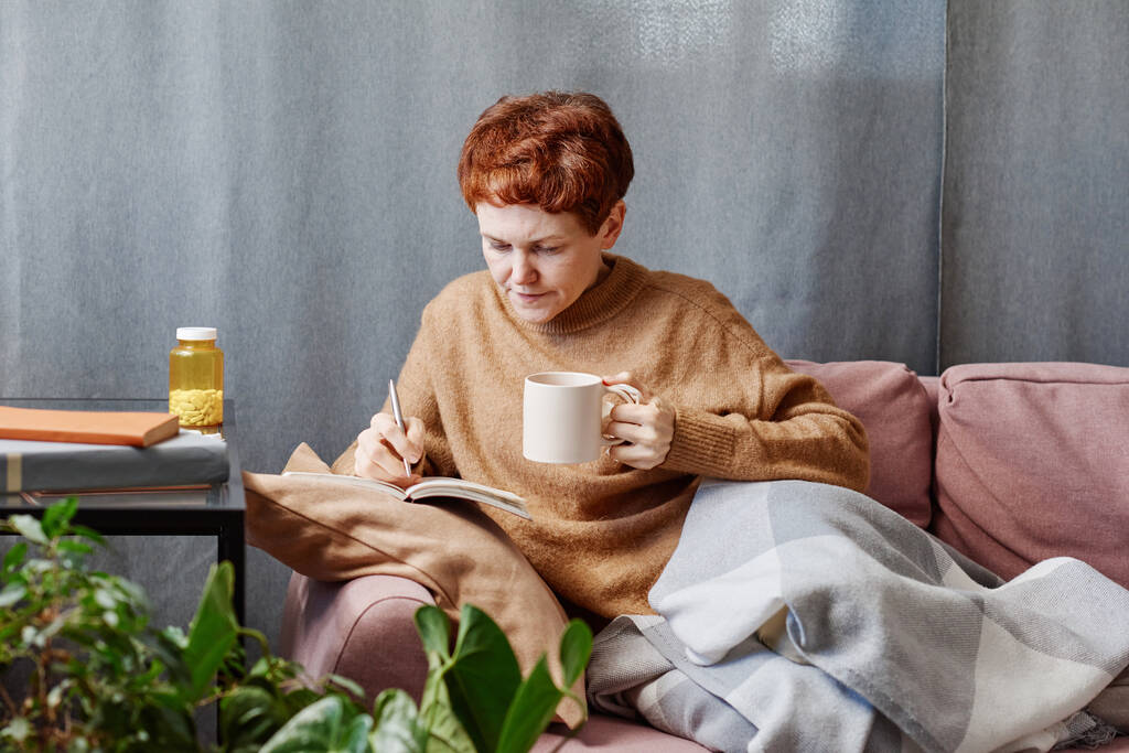 Vízszintes felvétel érett kaukázusi nőről, akinek az influenzája otthon marad, és jegyzetel valamit a jegyzetfüzetben és teát iszik. - Fotó, kép