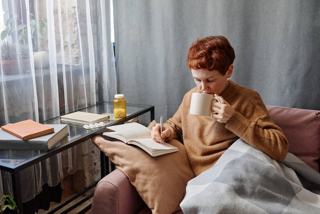 Υψηλής γωνίας πλάνο της σύγχρονης ώριμης Καυκάσιας γυναίκας που έχει γρίπη μένοντας στο σπίτι κάθεται στον καναπέ γράφοντας σημειώσεις στο σημειωματάριο και πίνοντας τσάι - Φωτογραφία, εικόνα