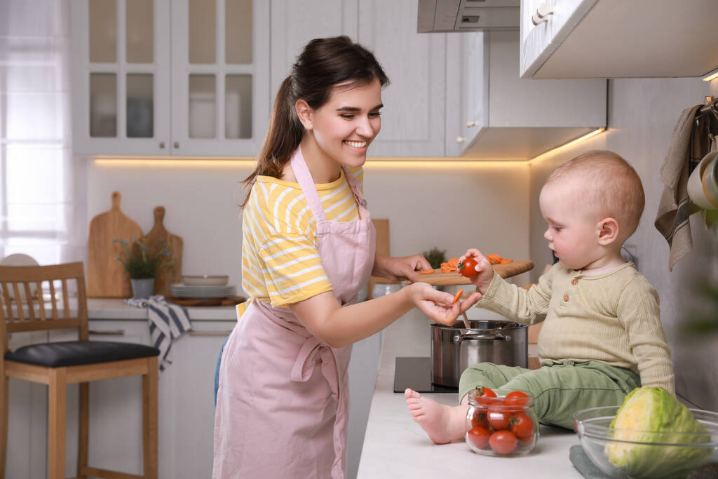 Ευτυχισμένη νεαρή γυναίκα με το χαριτωμένο μωρό της να περνάει χρόνο μαζί στην κουζίνα. - Φωτογραφία, εικόνα