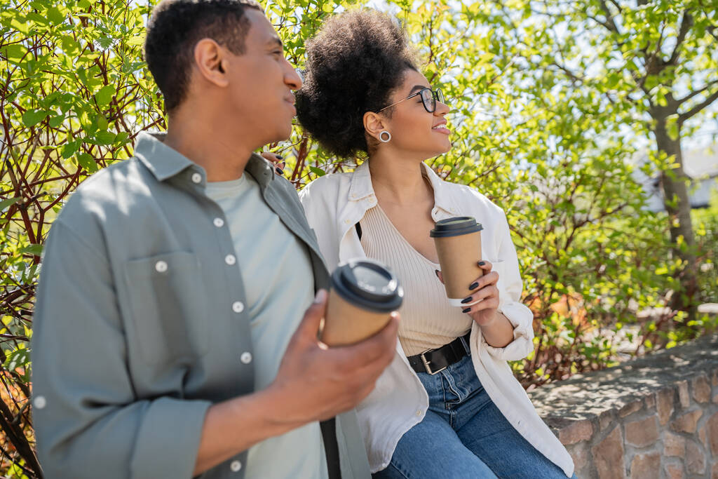 笑顔のアフリカ系アメリカ人女性がコーヒーを持って外でぼやけたボーイフレンドや木に近づく - 写真・画像