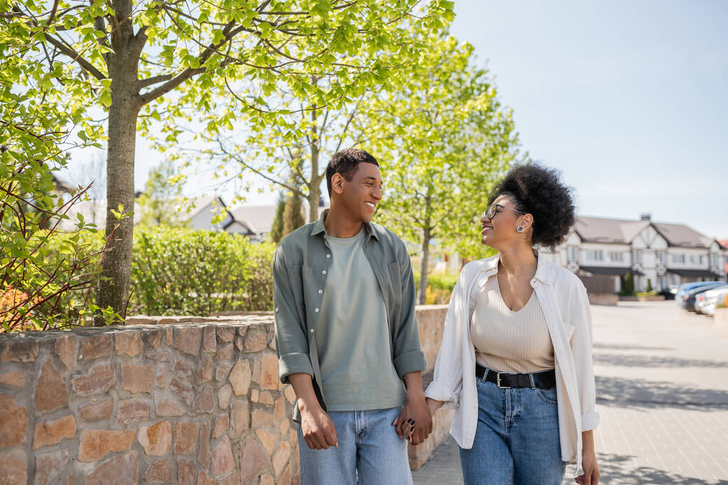 sorridente coppia afro-americana che si tiene per mano e parla mentre cammina sulla strada urbana in estate - Foto, immagini