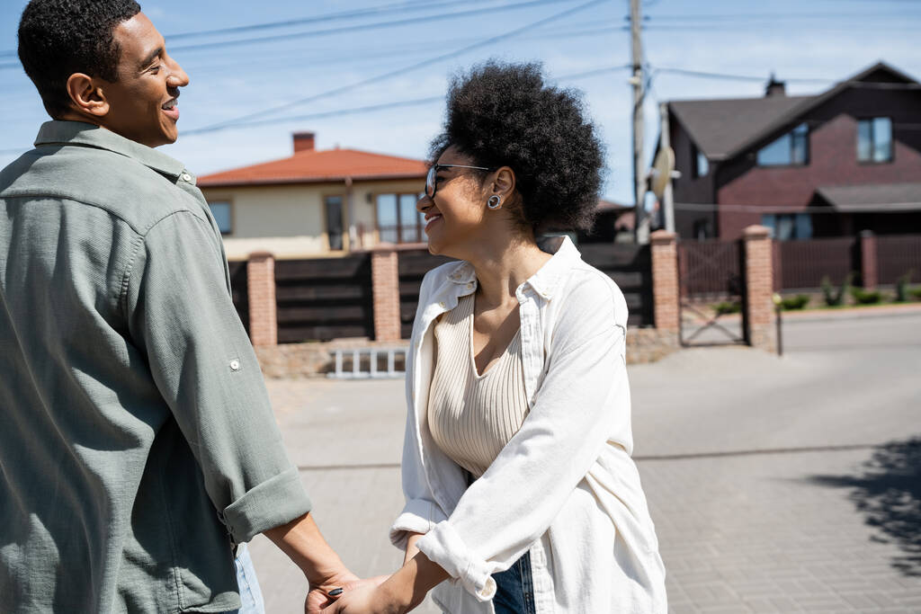 πλαϊνή άποψη του χαμογελαστού Αφροαμερικανού ζευγαριού κρατώντας τα χέρια σε αστικό δρόμο με τα σπίτια στο παρασκήνιο - Φωτογραφία, εικόνα