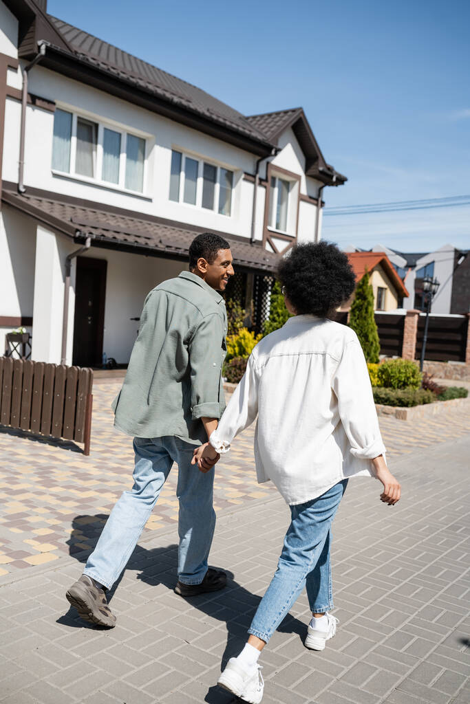 glimlachend Afrikaans Amerikaans paar hand in hand tijdens het lopen op de stoep in de buurt van huizen op straat - Foto, afbeelding