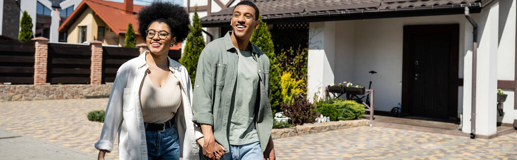 joyeux couple afro-américain tenant la main et marchant ensemble près des maisons dans la rue, bannière - Photo, image