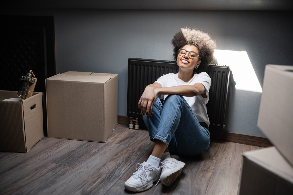 mulher americana africana feliz sentado à luz do sol perto de caixas de papelão no sótão na nova casa - Foto, Imagem