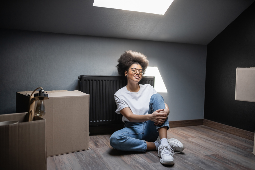 vrolijk Afrikaans amerikaanse vrouw in bril zitten in de buurt van kartonnen dozen op zolder in nieuw huis - Foto, afbeelding