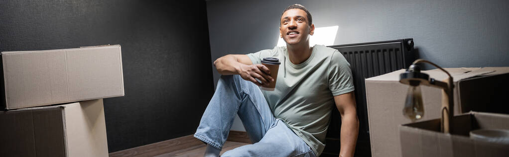 alegre afroamericano hombre sosteniendo café para ir cerca de cajas de cartón en casa nueva, pancarta - Foto, imagen