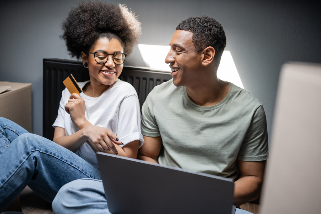 joyeux couple afro-américain en utilisant un ordinateur portable et une carte de crédit près de boîtes en carton dans une nouvelle maison - Photo, image