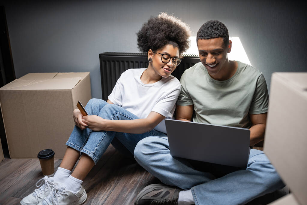 pareja afroamericana positiva usando el ordenador portátil y la tarjeta de crédito en el ático en la casa nueva - Foto, Imagen