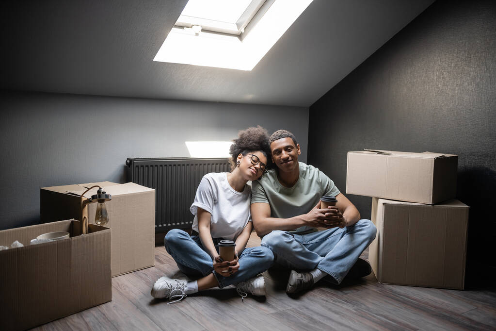 улыбающаяся африканская американская пара держит кофе, чтобы пойти, сидя рядом с коробками на чердаке в новом доме - Фото, изображение