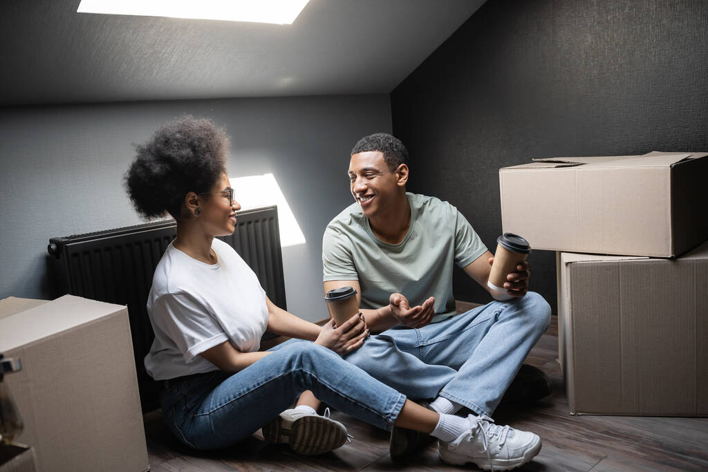 alegre africano americano pareja con café hablando cerca de cajas de cartón en ático en nueva casa - Foto, Imagen