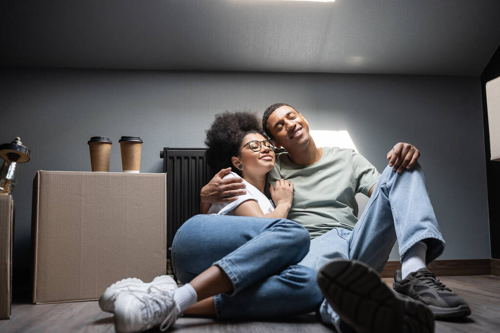 Neşeli Afrikalı Amerikalı çift, yeni evlerinde karton kutunun üstünde kahvenin yanında sarılıyorlar. - Fotoğraf, Görsel