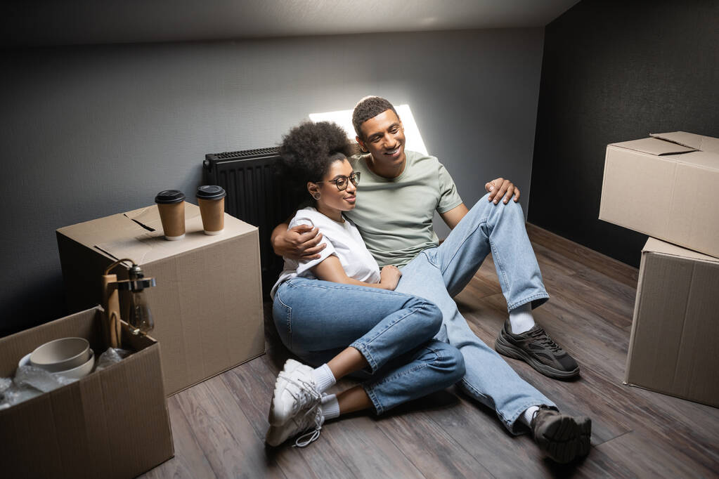 vreugdevol romantisch Afrikaans Amerikaans paar knuffelen in de buurt van koffie te gaan op dozen op zolder in nieuw huis - Foto, afbeelding