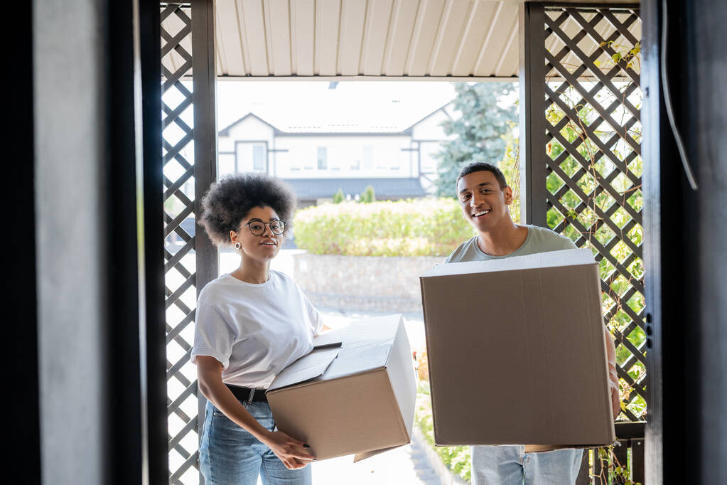 alegre africano americano pareja con paquetes mirando a cámara cerca puerta de nueva casa - Foto, imagen
