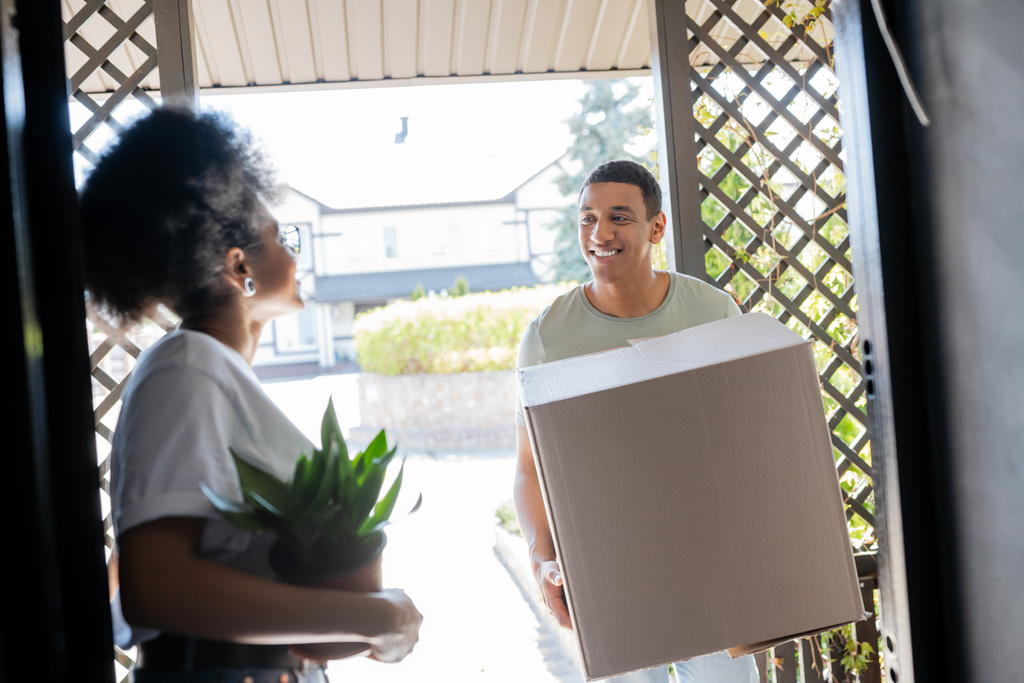 sonriente afroamericano hombre sosteniendo caja de cartón cerca de novia con planta en puerta de nueva casa - Foto, imagen