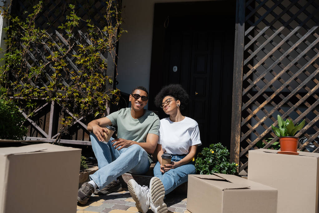 feliz africano americano pareja celebración de café y sentado cerca de paquetes y nueva casa - Foto, Imagen