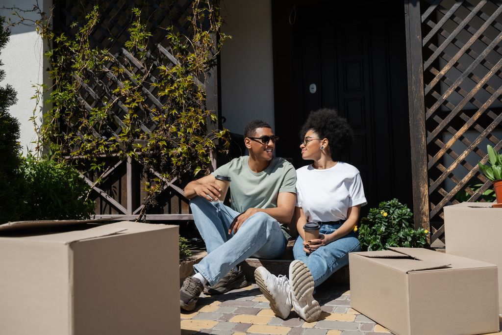 mosolygós afro-amerikai férfi napszemüvegben kezében kávé és beszél barátnőjével közelében új ház - Fotó, kép