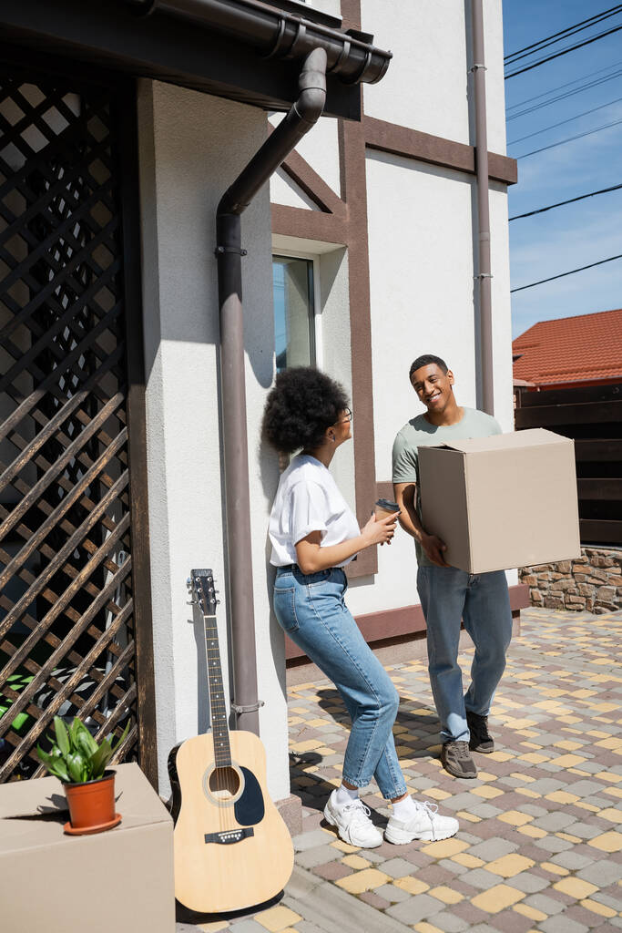 sonriente afroamericano hombre sosteniendo caja de cartón cerca de novia con café y casa nueva - Foto, Imagen