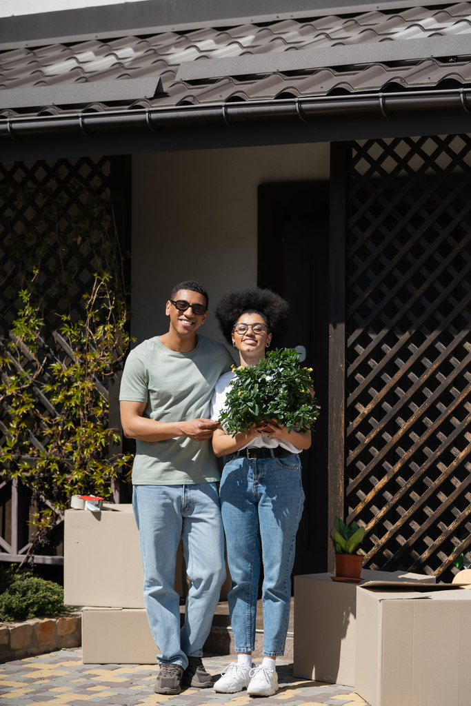 glückliches afrikanisch-amerikanisches Paar hält Zimmerpflanze in der Nähe von Paketen und neuem Haus im Freien - Foto, Bild