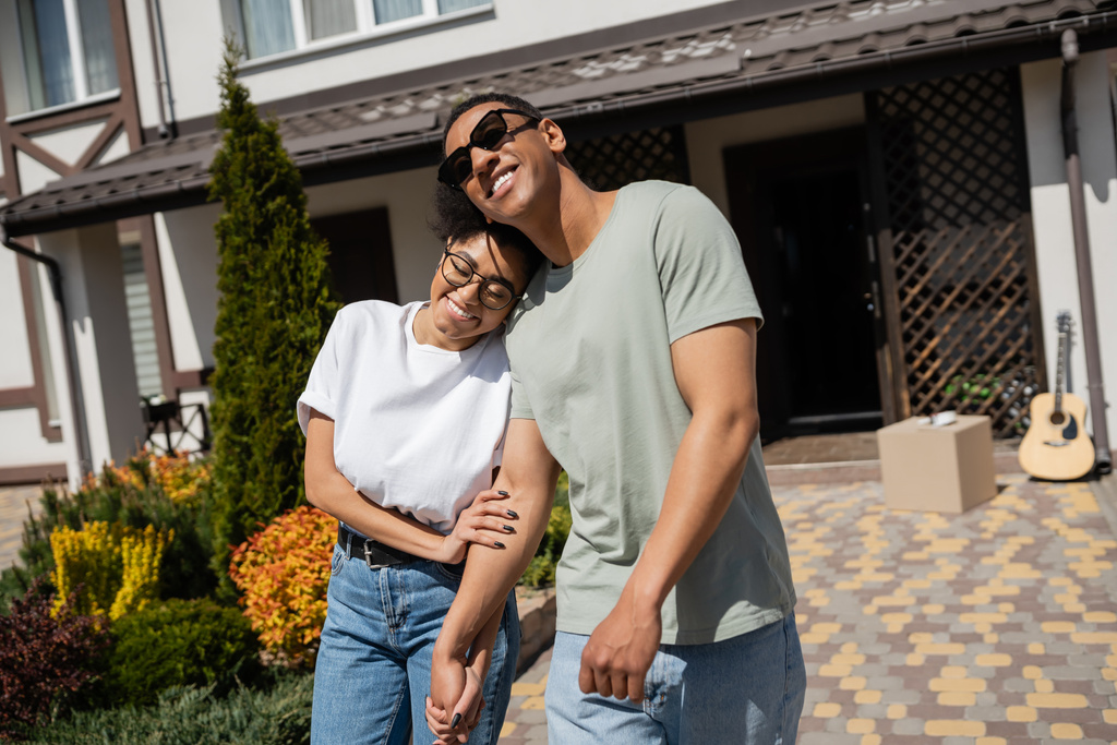 alegre casal afro-americano de mãos dadas enquanto está perto de nova casa no fundo ao ar livre - Foto, Imagem