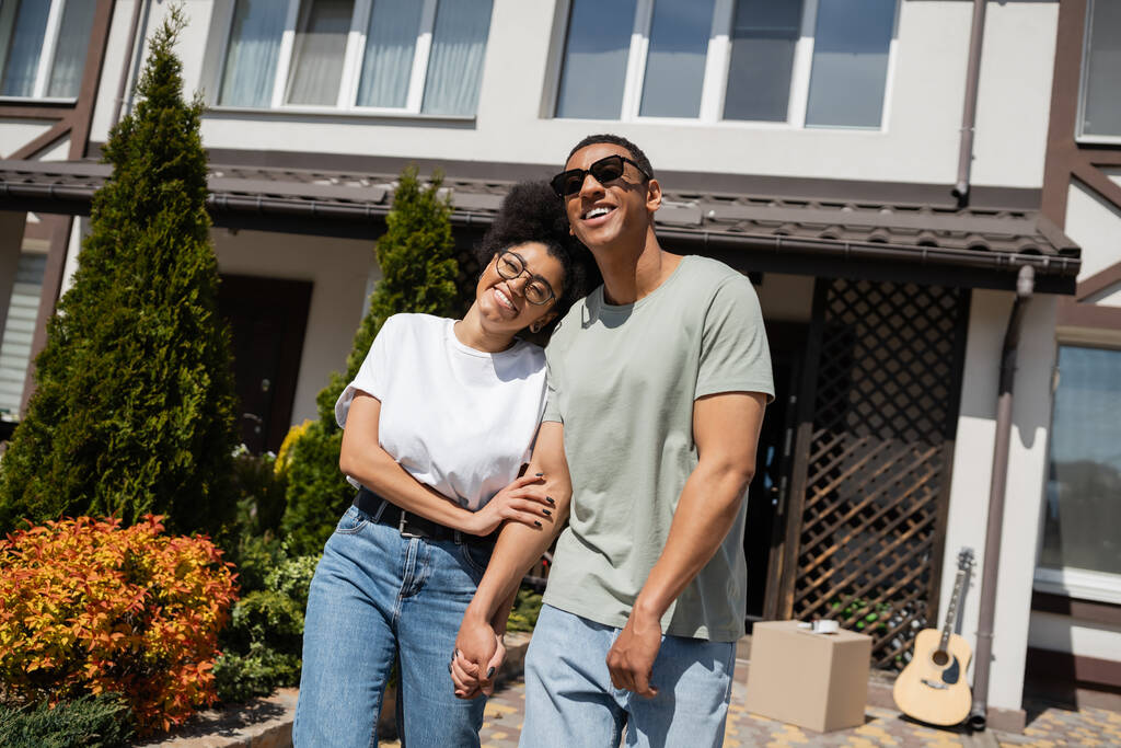 usmívající se africká americká žena v brýlích objímání přítel v blízkosti nového domu v pozadí - Fotografie, Obrázek