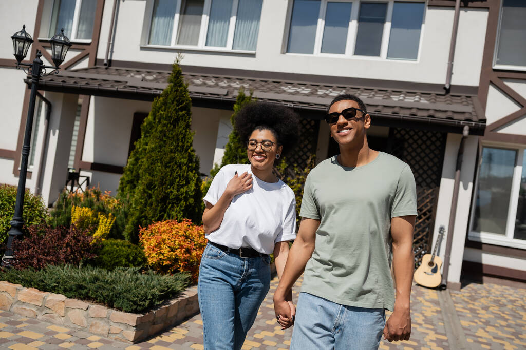 pozitivní romantický africký americký pár drží za ruce, zatímco stojí v blízkosti nového domu - Fotografie, Obrázek