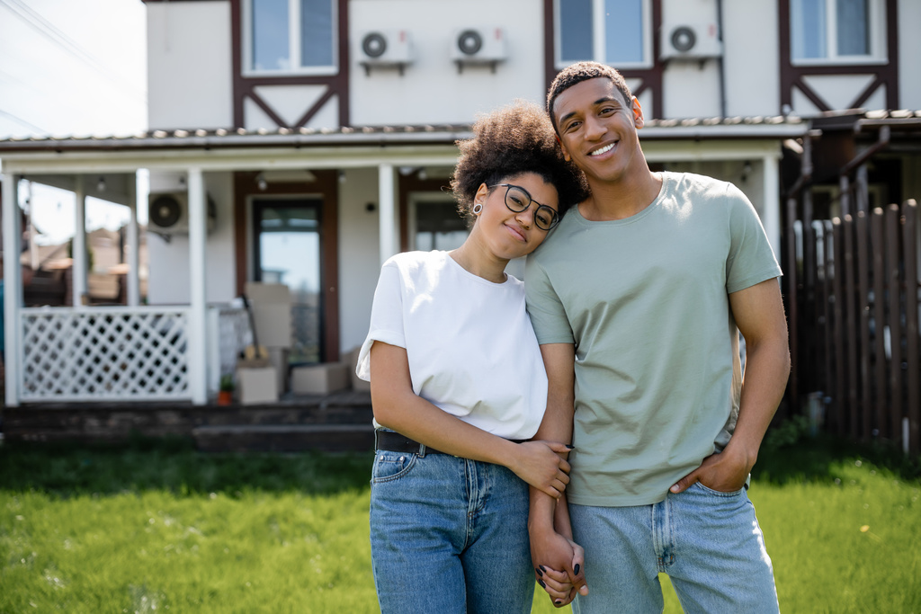 Mutlu Afro-Amerikan çifti el ele tutuşup arka plandaki yeni evin yakınındaki kameraya bakıyor. - Fotoğraf, Görsel