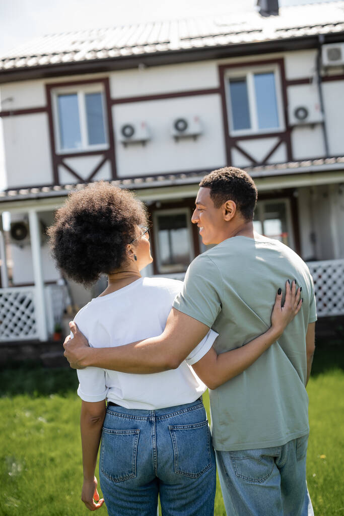 笑顔のアフリカ系アメリカ人の男がガールフレンドを抱きしめ,新しいぼやけた家の近くで話す - 写真・画像
