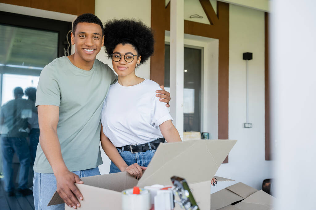 lächelnder afrikanisch-amerikanischer Mann umarmt Freundin in der Nähe von Kartons und neuem Haus während des Umzugs - Foto, Bild