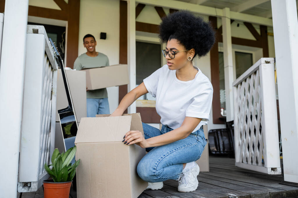Αφροαμερικανή γυναίκα ξεπακετάρει χαρτονένιο κουτί κοντά στο θολή φίλο και το νέο σπίτι - Φωτογραφία, εικόνα